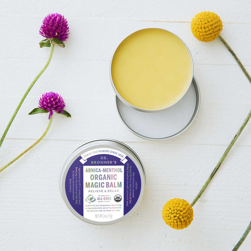 An open tin of Dr. Bronners Arnica Menthol Organic Magic Balm sits on a white surface, surrounded by colorful flowers. The label reads Relieve Relax, and All One with organic certification symbols  [2 oz]