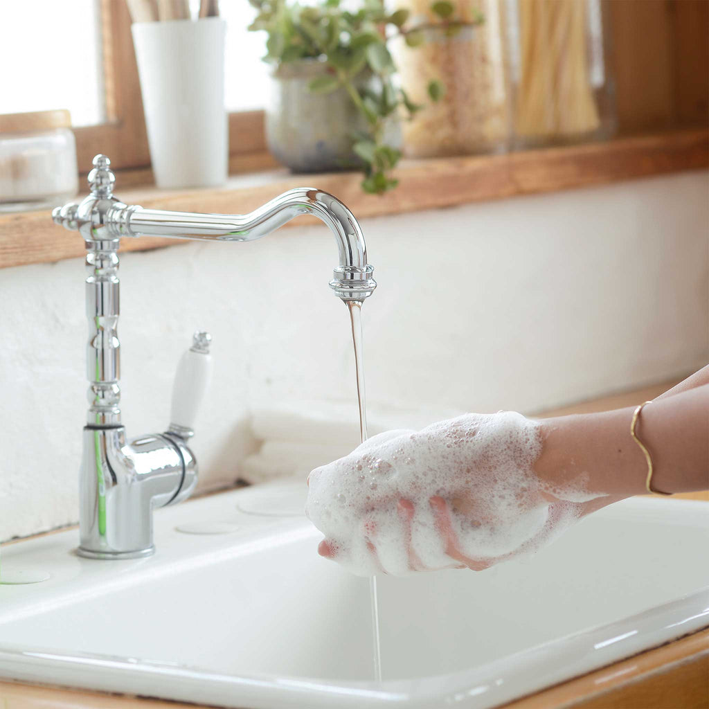 A hand lathers with foamy soap under a sleek chrome faucet, set in a white ceramic sink. Potted plants and light filtering through a window create a serene, natural environment [12 oz, 24 oz, 1/2 Gallon]