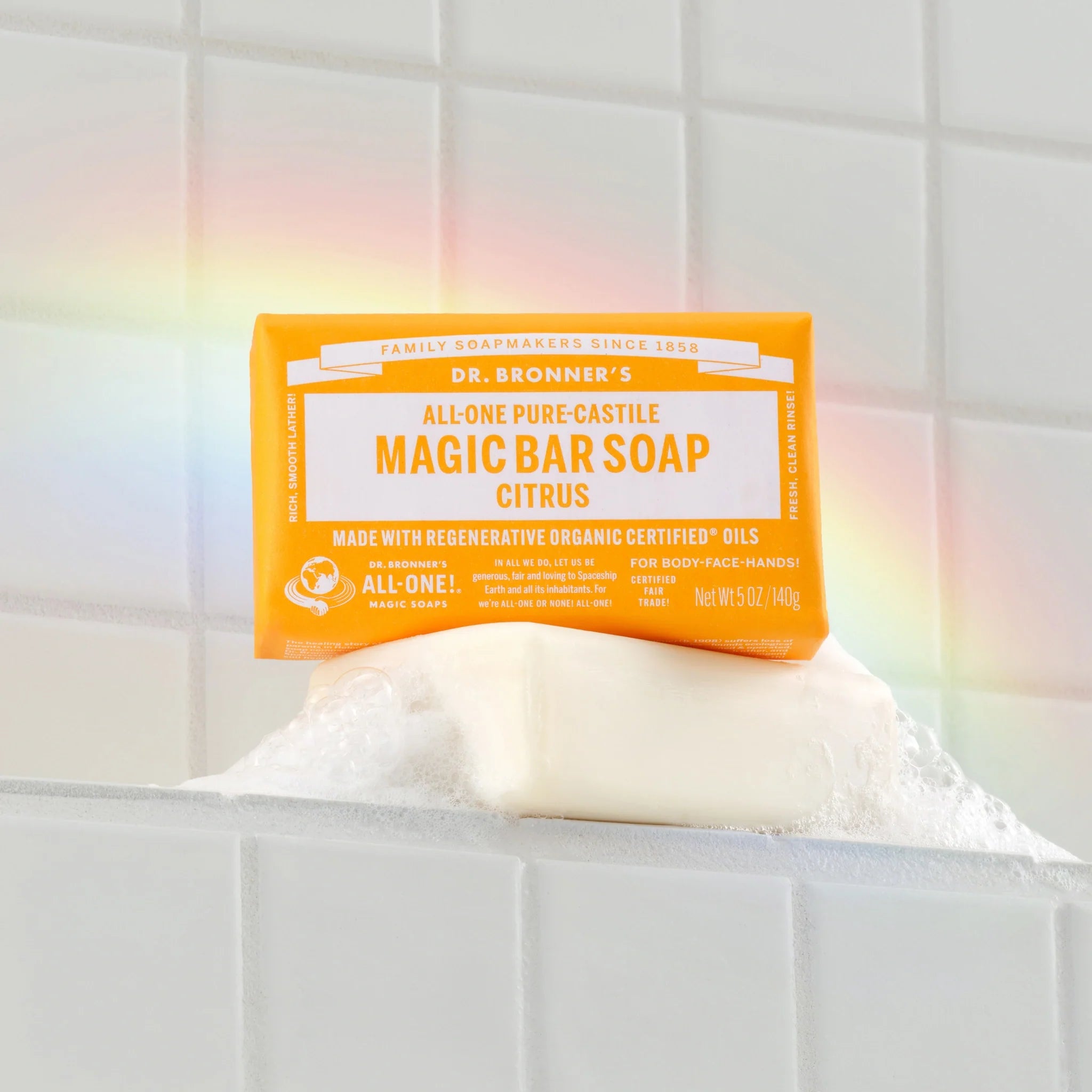 Dr. Bronners citrus magic bar soap rests on a foamy white tile surface with a subtle rainbow glow. All One Pure Castile Magic Bar Soap, Citrus, Made with Regenerative Organic Certified Oils, Net Wt 5 oz140 g [5 oz]