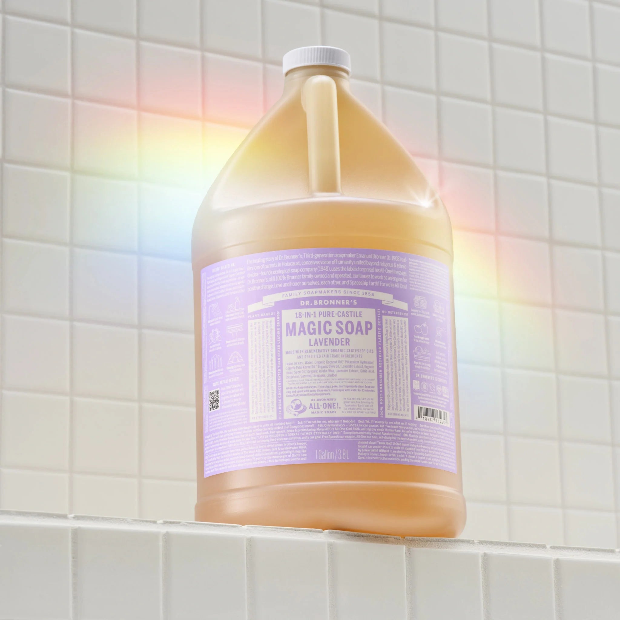 A large bottle of Dr. Bronners Lavender Pure Castile Soap sits on a tiled bathroom shelf, reflecting a rainbow light. The lavender label details its vegan, organic, and versatile cleaning uses [1 Gallon]