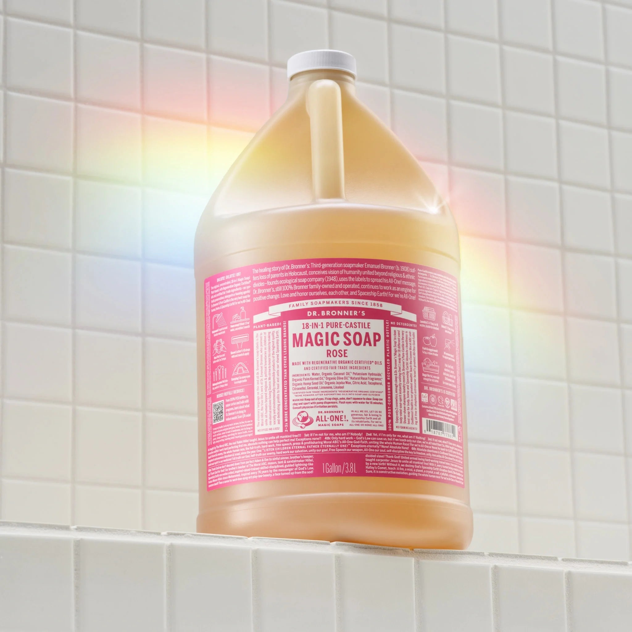 A gallon sized container of Dr. Bronners 18 in 1 Pure Castile Rose Magic Soap stands against a tiled wall, emitting a rainbow reflection. The label highlights its versatile, biodegradable, and organic nature [1 Gallon]