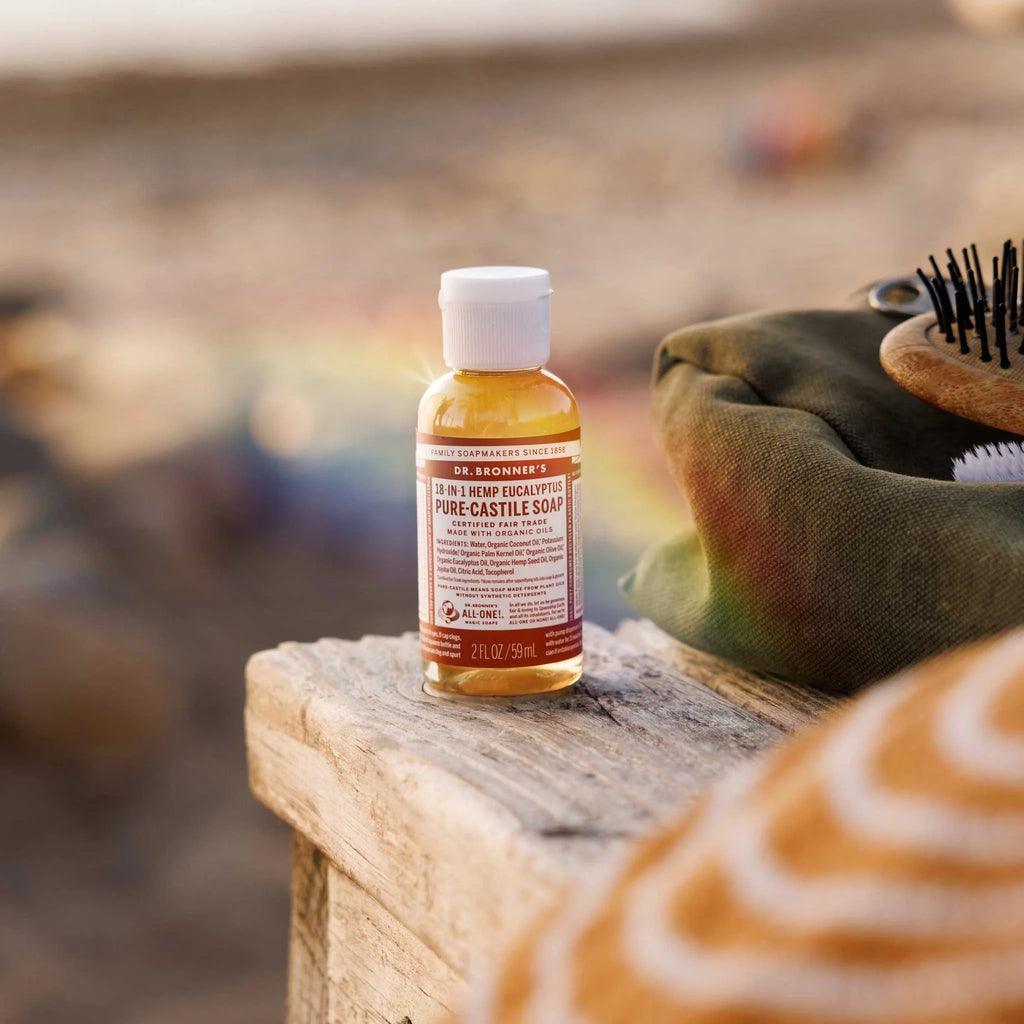 A small bottle of Dr. Bronners liquid soap sits on a wooden surface outdoors. The background features a blurred beach scene, creating a relaxed and natural context for this versatile, eco friendly product [2 oz]