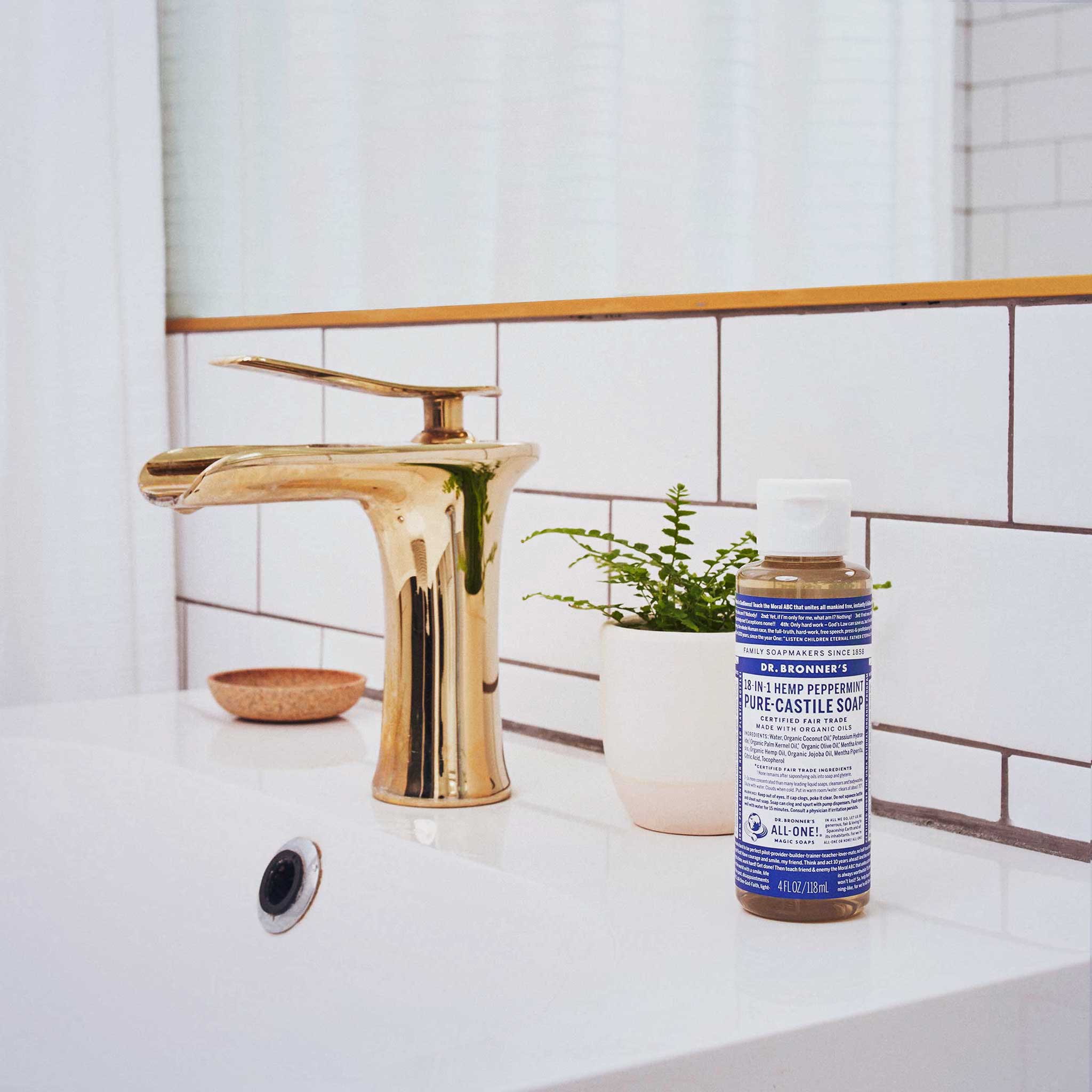A bottle of Dr. Bronners 18 in 1 Hemp Peppermint Pure Castile Soap sits on a bathroom counter beside a gold faucet and a small potted plant, against tiled walls [4 oz]