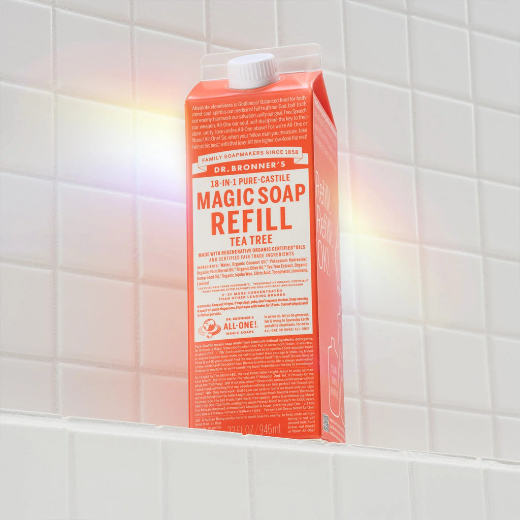 Dr. Bronners 18 in 1 Pure Castile Magic Soap Refill in Tea Tree scent sits upright on a white tiled shelf. The packaging highlights eco friendly and fair trade qualities, emphasizing sustainable practices [32 oz]