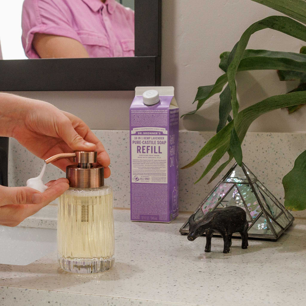 A hand pumps liquid soap from a dispenser on a bathroom counter, flanked by Dr Bronners lavender castile soap carton, a plant, and decorative item. Reflection visible in mirror [32 oz]