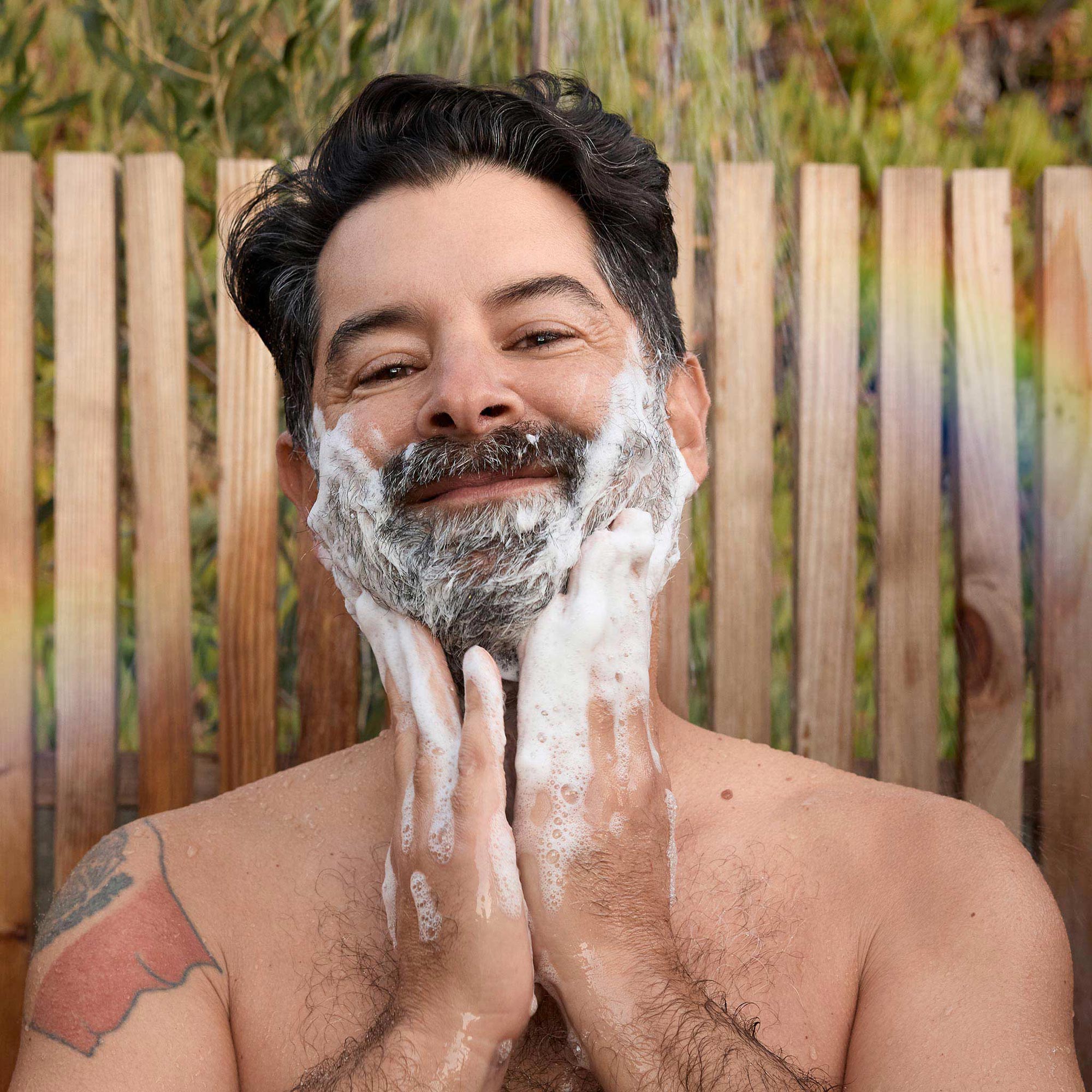 A man lathers his face with foamy dr bronners castile soap, smiling contentedly. He stands outdoors, in front of a wooden fence with lush greenery in the background, embracing an eco friendly, natural skincare routine [2 oz, 3.4 oz, 4 oz, 8 oz, 16 oz, 32 oz, 1/2 Gallon, 1 Gallon]